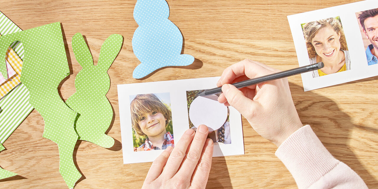 Dvě ruce kreslí kruh kolem hlavy člena rodiny pomocí šablony. Vedle ní jsou další vytištěné fotografie a vystřižení velikonoční zajíčci z barevného papíru.