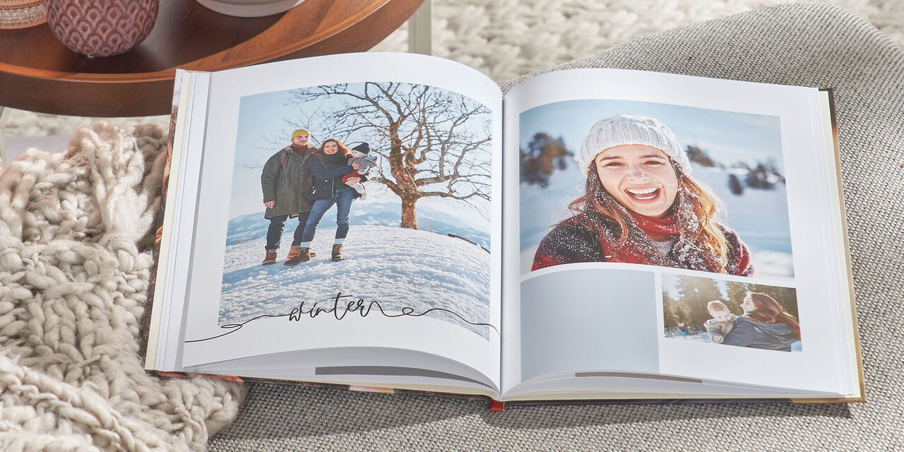 Na šedé pohovce leží otevřená CEWE FOTOKNIHA. Obsahuje několik fotografií rodiny a slovo "Winter". Vlevo je stolek s čajovými svíčkami a rostlinou.